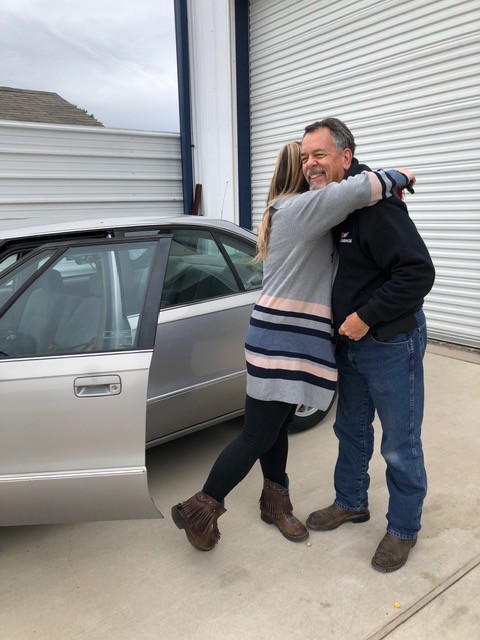 Man in God’s Garage hoodie hugging a woman beside an open car door, holding keys