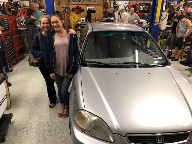 Two women smiling beside a silver Honda on a light gray flake epoxy floor inside God’s Garage